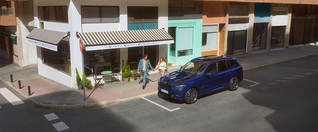 Paar geht Hand in Hand an einem geparkten blauen BMW X3 vor einem Café vorbei.
