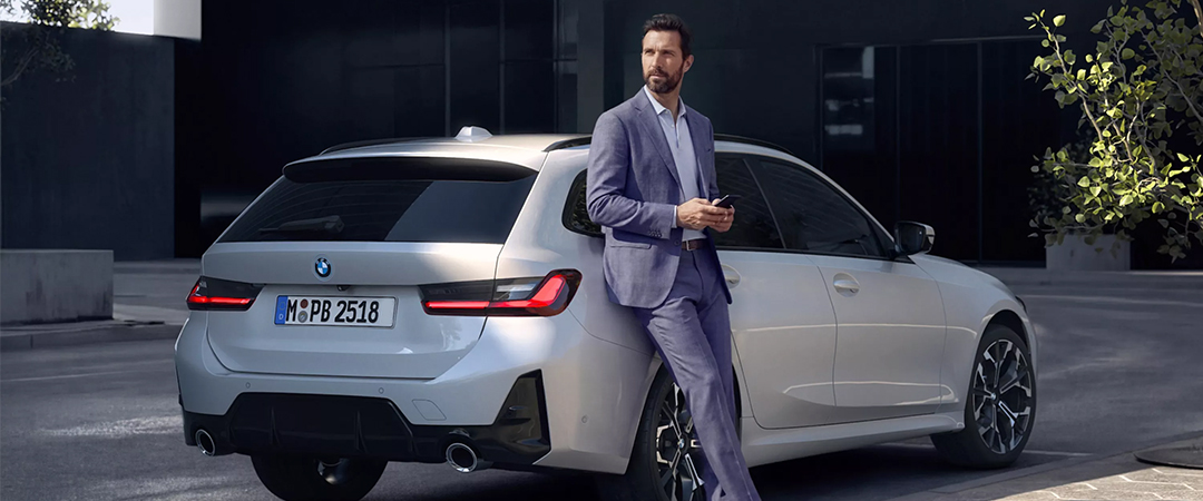 Mann im Anzug steht mit Smartphone neben einem weißen BMW 3er Touring.