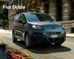 Ein grauer Fiat Doblo fährt auf einer sonnigen städtischen Straße entlang. Im Vordergrund sind leicht verschwommene Blumen zu sehen, während im Hintergrund historische Gebäude stehen. Oben links im Bild steht der Text „Fiat Doblo“.