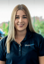 Brustbild einer jungen, lächelnden Frau mit langen, blonden Haaren. Sie trägt ein dunkelblaues Poloshirt mit dem weißen Logo "RheinRent" auf der rechten Brust. Der Hintergrund ist hell und unscharf.
