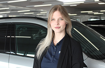Dieses Bild zeigt ebenfalls eine junge, lächelnde Frau im Nahbereich. Sie hat langes, sehr hellblondes Haar und trägt einen kleinen Nasenring. Auch sie trägt ein weißes Poloshirt mit Kragen, auf dem ein dunkles Logo zu sehen ist. Sie steht vor einem hellen Auto, das im Hintergrund unscharf erkennbar ist, was auf ein Autohaus oder einen Verkaufsraum hindeutet. Das Licht ist hell und natürlich.