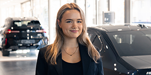 Juliana Lange - Assistenz Leitung Rhein Rent Das Bild zeigt eine junge, lächelnde Frau mit schulterlangem, blondem Haar. Sie trägt einen schwarzen Blazer über einem schwarzen Oberteil und eine zarte Halskette. Sie steht in einem Autohaus, erkennbar an den neuen, dunklen Autos im unscharfen Hintergrund links und rechts von ihr. Die Beleuchtung ist hell und der Fokus liegt klar auf der Frau.