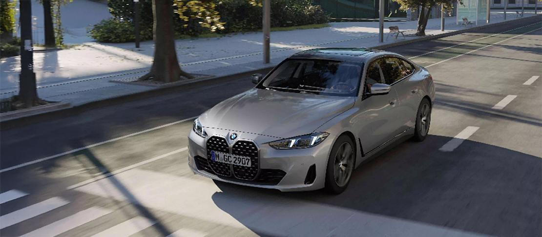  Ein Bild, das den silbernen BMW 4er Gran Coupé im leichten Schrägblick von vorne zeigt, wie er auf einer asphaltierten Straße fährt. Die Umgebung ist eine städtische Allee mit Bäumen, Gehwegen und einer Bushaltestelle. Das Auto ist in Bewegung, was durch leichte Unschärfe des Hintergrunds angedeutet wird.