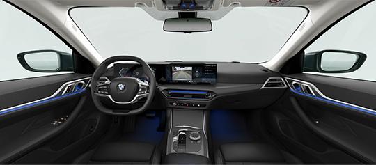 Eine Aufnahme des Innenraums des BMW 4er Gran Coupé, die die Fahrerkabine aus der Mitte der Rückbank zeigt. Zu sehen sind das Armaturenbrett mit dem großen Curved Display, das Lenkrad, die Mittelkonsole und die Sitze. Blaue Akzentbeleuchtung (Ambient Light) ist an den Türen und im Fußraum sichtbar.