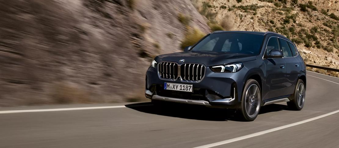  Ein Bild, das den dunkelblauen BMW X1 SUV im leichten Schrägblick von vorne zeigt, wie er dynamisch eine kurvige Bergstraße entlangfährt. Im Hintergrund ist eine felsige, trockene Landschaft zu sehen. Das Auto ist in Bewegung.
