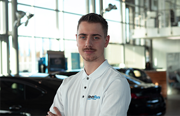 Das Bild zeigt einen jungen Mann mit kurzen, dunklen Haaren und einem leichten Bart, der in die Kamera blickt. Er trägt ein weißes Poloshirt mit einem Firmenlogo auf der Brust und hat die Arme vor dem Körper verschränkt. Er steht in einem hellen, modernen Autohaus oder Showroom, erkennbar an den großen Fensterfronten und den unscharf im Hintergrund sichtbaren dunklen Fahrzeugen.