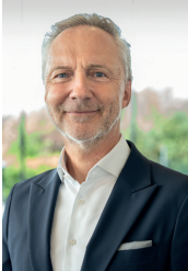 Porträtfoto eines grauhaarigen Mannes mittleren Alters, der freundlich lächelt und einen dunklen Anzug mit einem weißen Hemd trägt. Er trägt ein Einstecktuch und steht vor einem hellen, leicht verschwommenen Hintergrund.