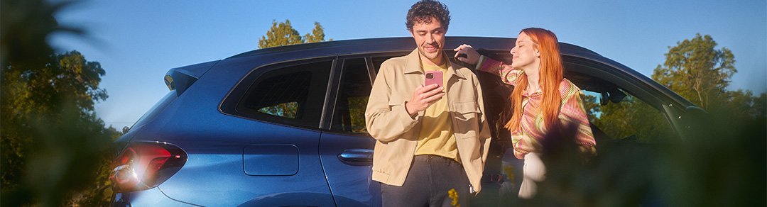 Paar steht neben einem blauen BMW und schaut gemeinsam auf ein Smartphone.