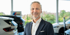 Ralph Klapper - Leitung Rhein Rent Dieses Bild zeigt einen älteren Mann mit kurzen, grau melierten Haaren und einem gepflegten Bartansatz, der freundlich lächelt und direkt in die Kamera blickt. Er trägt einen dunkelblauen Anzug mit einem weißen Einstecktuch und einem weißen Hemd ohne Krawatte. Er steht in einem modernen Autohaus, erkennbar an den hellen Autos und einem dezenten Logo im unscharfen Hintergrund links. Große Fenster im Hintergrund lassen viel Tageslicht herein.