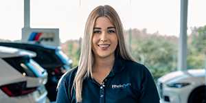 Viktoria Hörlin - Managerin Rhein Rent Dieses Bild zeigt eine junge, lächelnde Frau mit langem, glattem Haar, das oben dunkel und zu den Spitzen hin heller (Ombre) wird. Sie trägt ein dunkelblaues Poloshirt mit einem hellen Logo auf der Brust. Sie steht in einem modernen Autohaus. Im unscharfen Hintergrund sind weiße und dunkle Autos erkennbar. Links ist ein dezentes rotes, blaues und weißes Logo an der Wand zu sehen, und große Fenster lassen helles Tageslicht in den Raum.